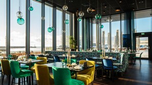 Un restaurante con amplios ventanales, sillas verdes y amarillas, y lámparas esféricas colgantes.