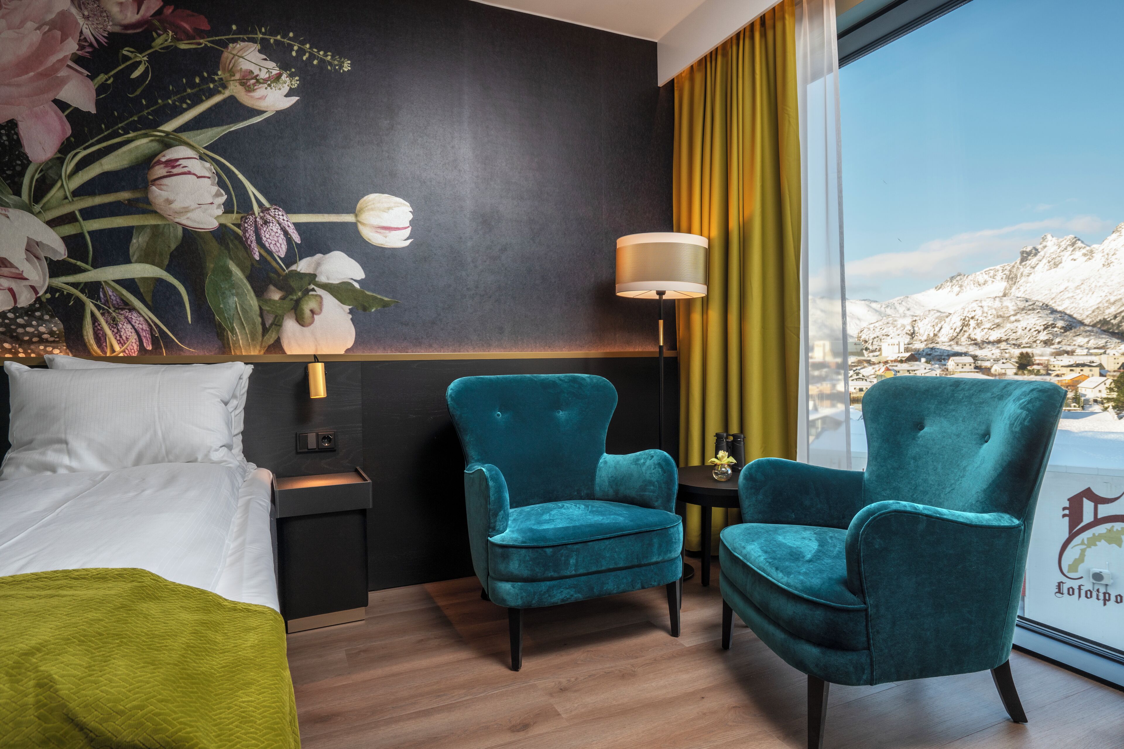 A hotel room with a bed, two teal armchairs, a nightstand with a lamp, and a large window with a view of snow-capped mountains.