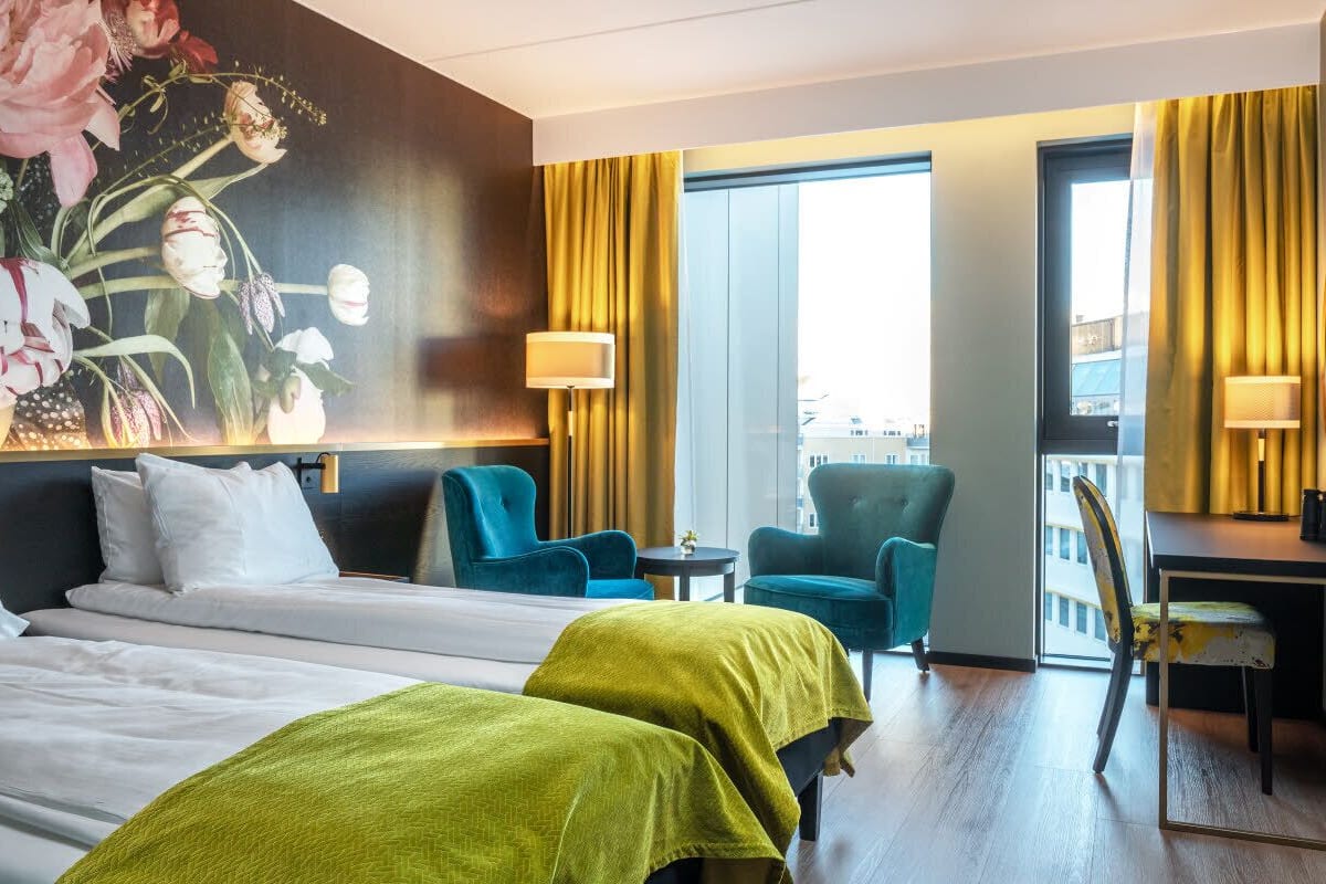 Quarto de hotel com duas camas, uma escrivaninha, uma cadeira e um grande mural floral na parede.