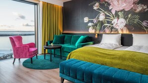 Habitación de hotel moderna con una cama grande, un sofá verde, un sillón rosa y vistas al mar a través de un ventanal amplio.