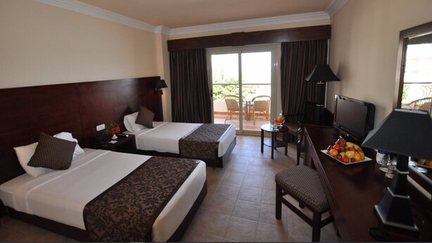 A hotel room with two beds, a desk with a television, a dining table with fruit, and a balcony with outdoor furniture. - Royal Brayka Beach Resort (Marsa Alam)
