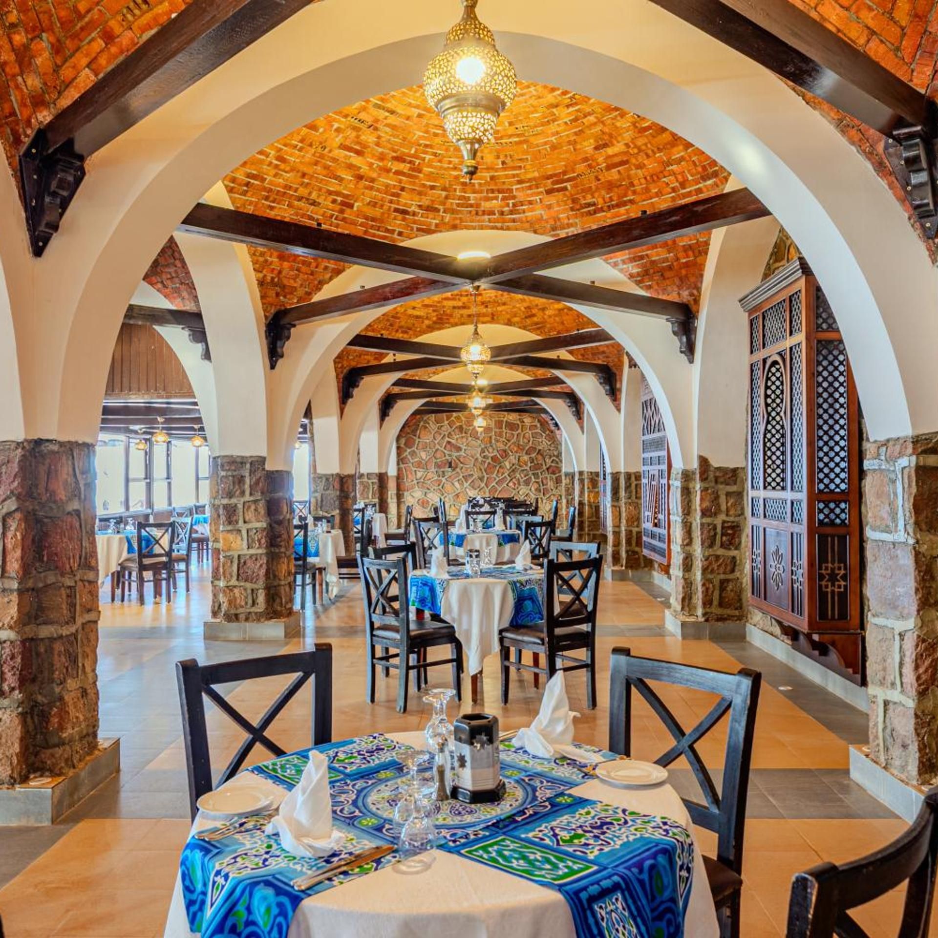 Restaurante com teto de tijolos, paredes de pedra e toalha de mesa estampada. Mesas montadas com toalhas brancas e talheres.