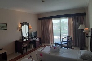 A hotel room with a bed, a desk with a computer, a chair, a large window with curtains, and a view of the outdoors. - Royal Brayka Beach Resort (Marsa Alam)