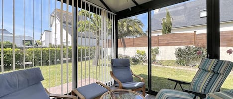 Une terrasse ensoleillée, protégée par une structure en verre, aménagée avec des fauteuils en osier et une table en verre, et offrant une vue sur un quartier résidentiel.
