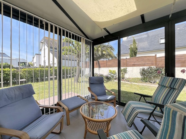 Une terrasse ensoleillée, protégée par une structure en verre, aménagée avec des fauteuils en osier et une table en verre, et offrant une vue sur un quartier résidentiel.