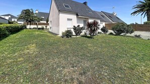 Una casa residencial con techo inclinado, un pequeño jardín delantero con césped y arbustos, y un cielo azul despejado.