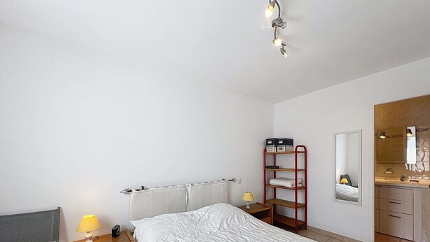 Habitación de hotel con cama, lámparas de noche, mesita de noche de madera, estante metálico rojo, espejo y baño con lavamanos y espejo.