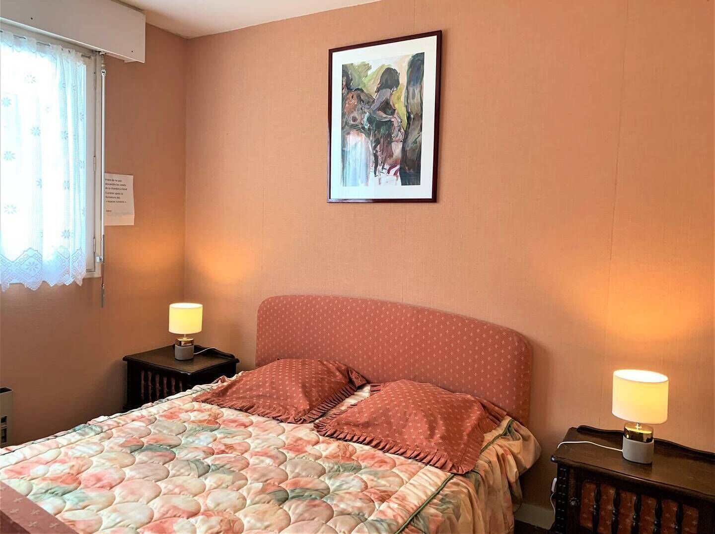 A bedroom with a bed, two bedside tables with lamps, a framed picture on the wall, and a window with a curtain.