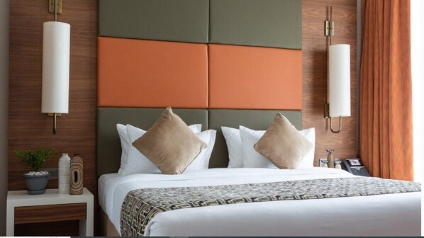 A hotel room with a bed, a nightstand with a plant and decorative items, and a wall-mounted lamp. - Emara Ole-sereni (Nairobi)