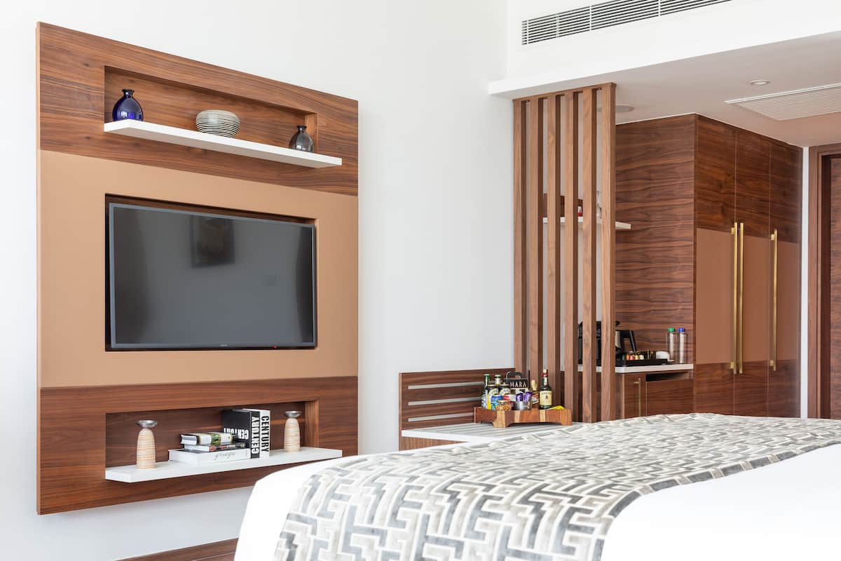 Un dormitorio moderno con un televisor de pantalla plana grande montado en la pared, estanterías de madera con objetos decorativos, una cama con cubrecama estampado y un armario con puerta espejada.