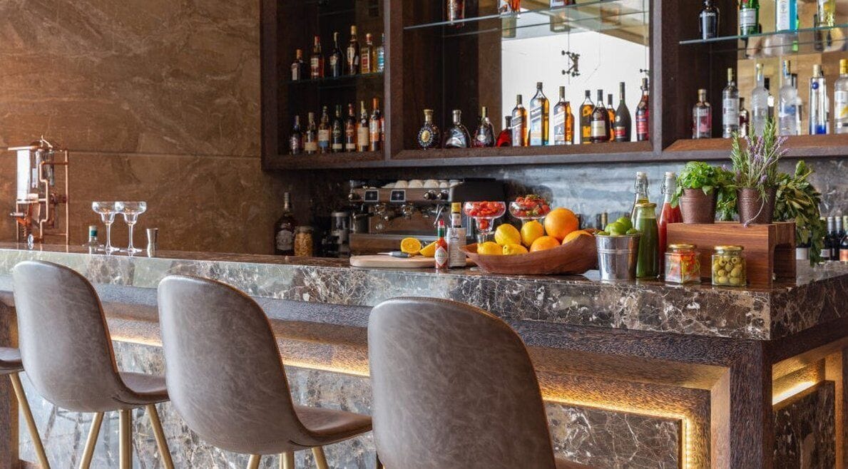 Una zona de bar con encimera de mármol, taburetes y una variedad de botellas y vasos.