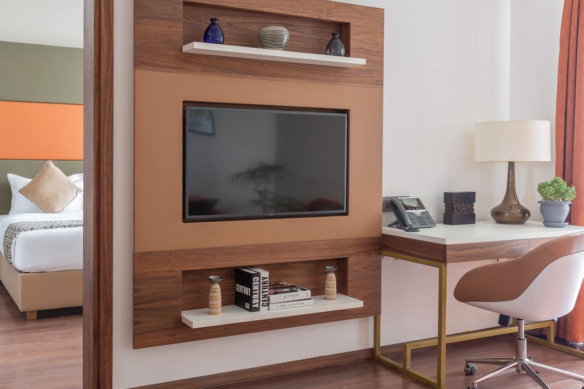 Habitación de hotel moderna con un televisor de pantalla plana grande montado en una pared de madera, un escritorio con lámpara, una silla y una mesita de noche con una planta.