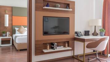 A modern hotel room with a large flat-screen TV mounted on a wooden wall, a desk with a lamp, a chair, and a bedside table with a plant.