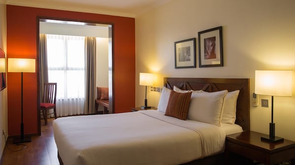 Habitación de hotel con una cama grande, dos lámparas de noche, una mesita de noche, una silla, una ventana con cortinas y dos cuadros en la pared.