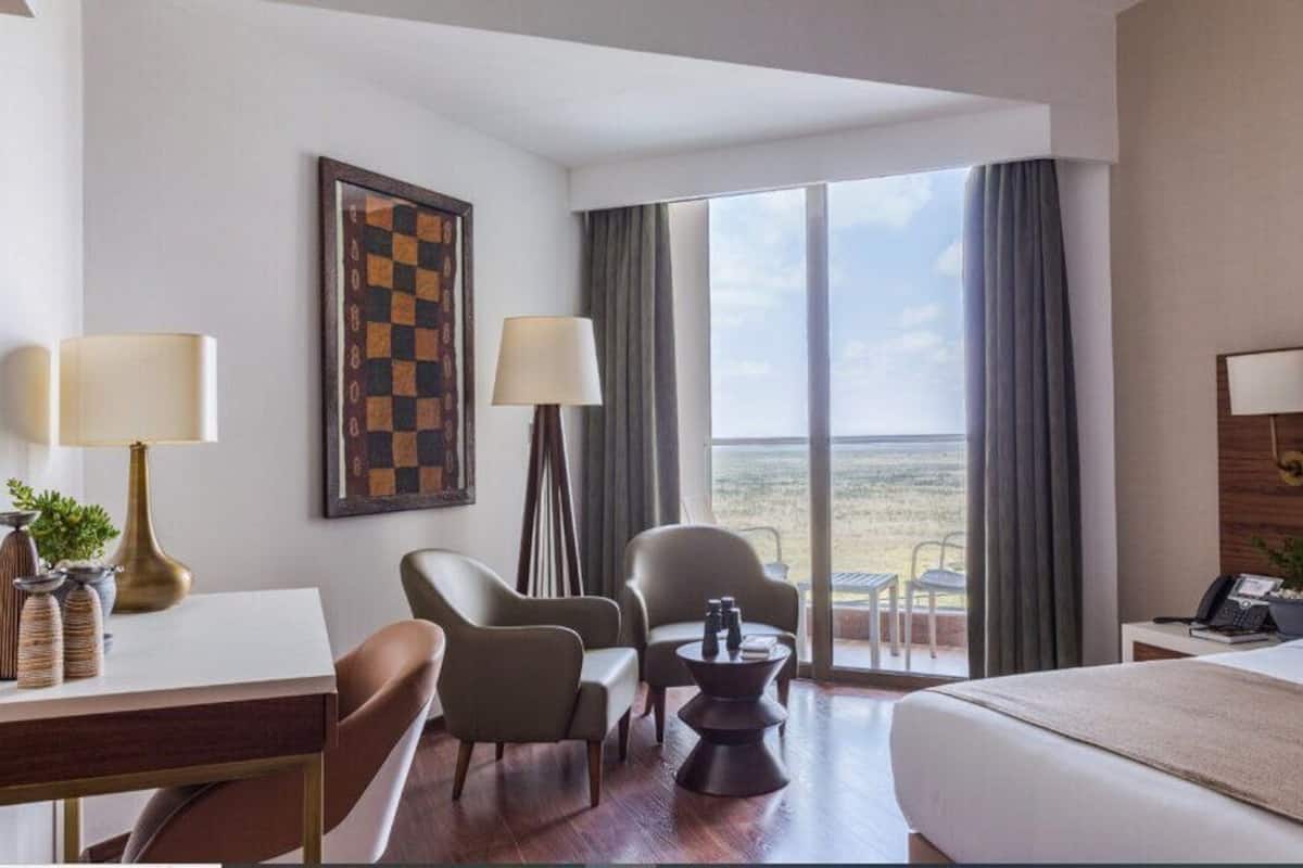 Habitación de hotel con cama, dos sillas, escritorio, lámpara, un tablero de ajedrez enmarcado y vista a la playa.