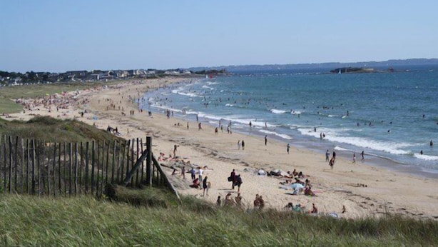 Una playa con valla de madera, orilla arenosa y gente disfrutando del agua.