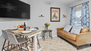 Ein Essbereich mit Tisch für zwei Personen, einer Couch, einem an der Wand montierten Fernseher und einem Fenster mit gemusterten Vorhängen.