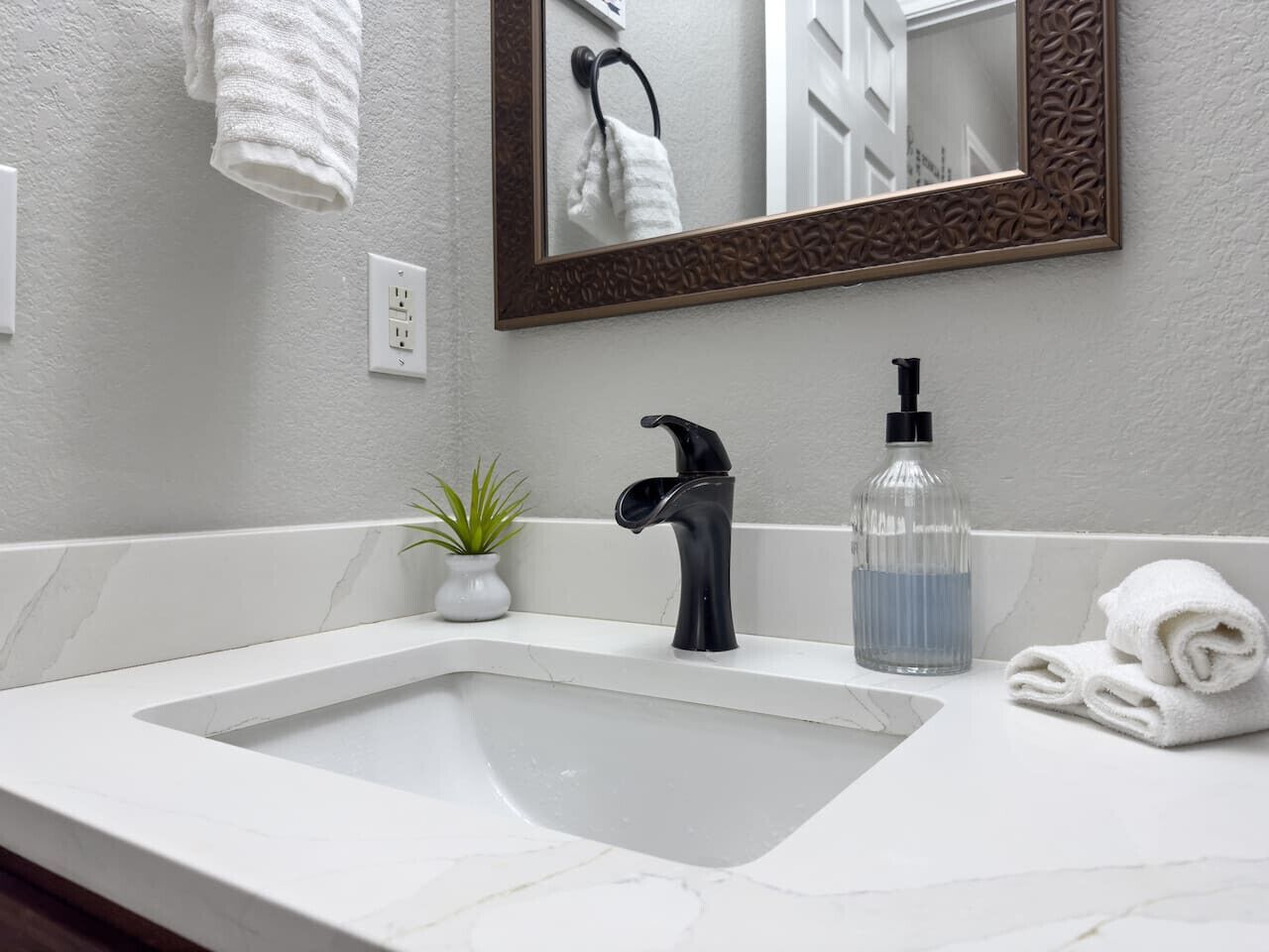 A bathroom with a white sink, black faucet, and a mirror with a decorative frame. There is a small potted plant and a bottle of soap on the counter.