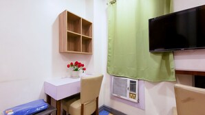 Una habitación pequeña con escritorio, silla y televisor. Hay un estante sobre el escritorio y una cortina en la ventana.