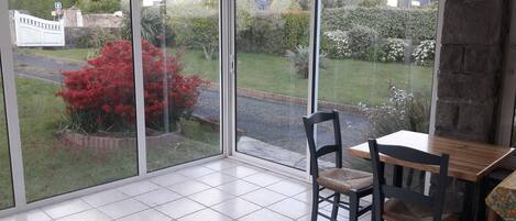 Une pièce dotée d’une grande porte vitrée donnant sur un jardin. À l’intérieur, on trouve une table en bois et deux chaises.