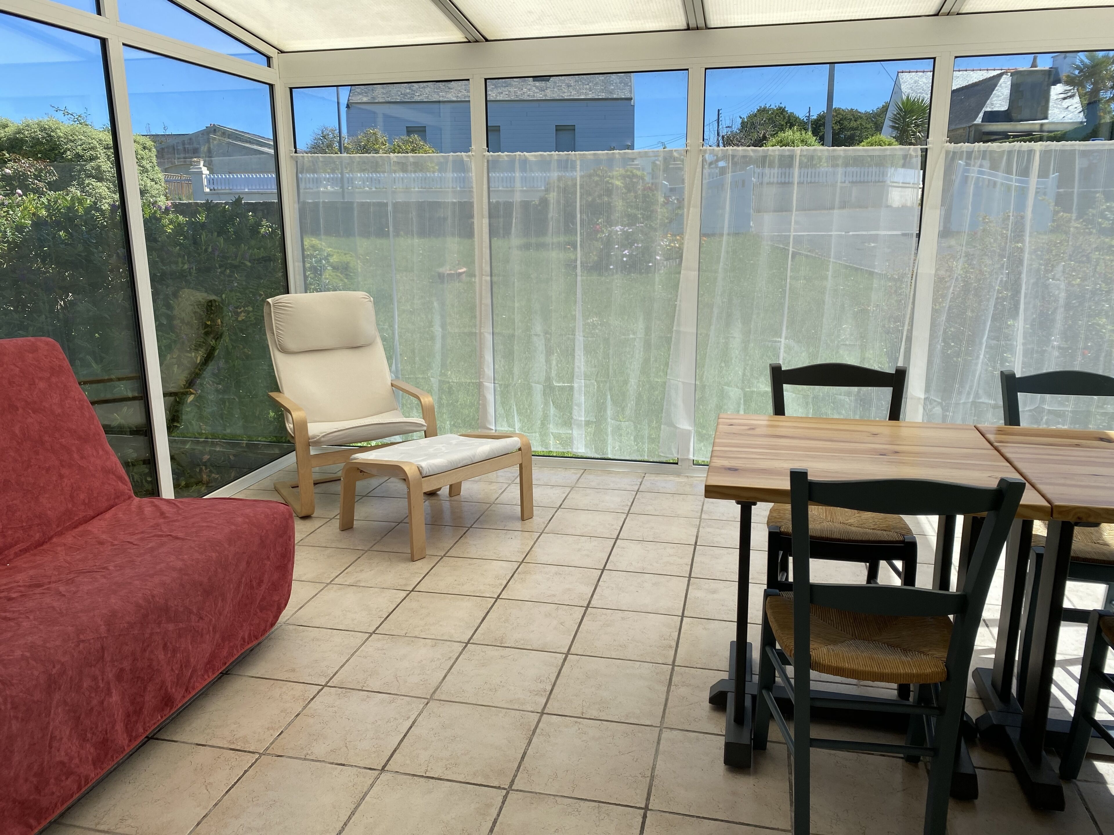 Een zonnedoorspoelde kamer met een rode bank, een houten stoel, een kleine tafel en een eettafel met stoelen.