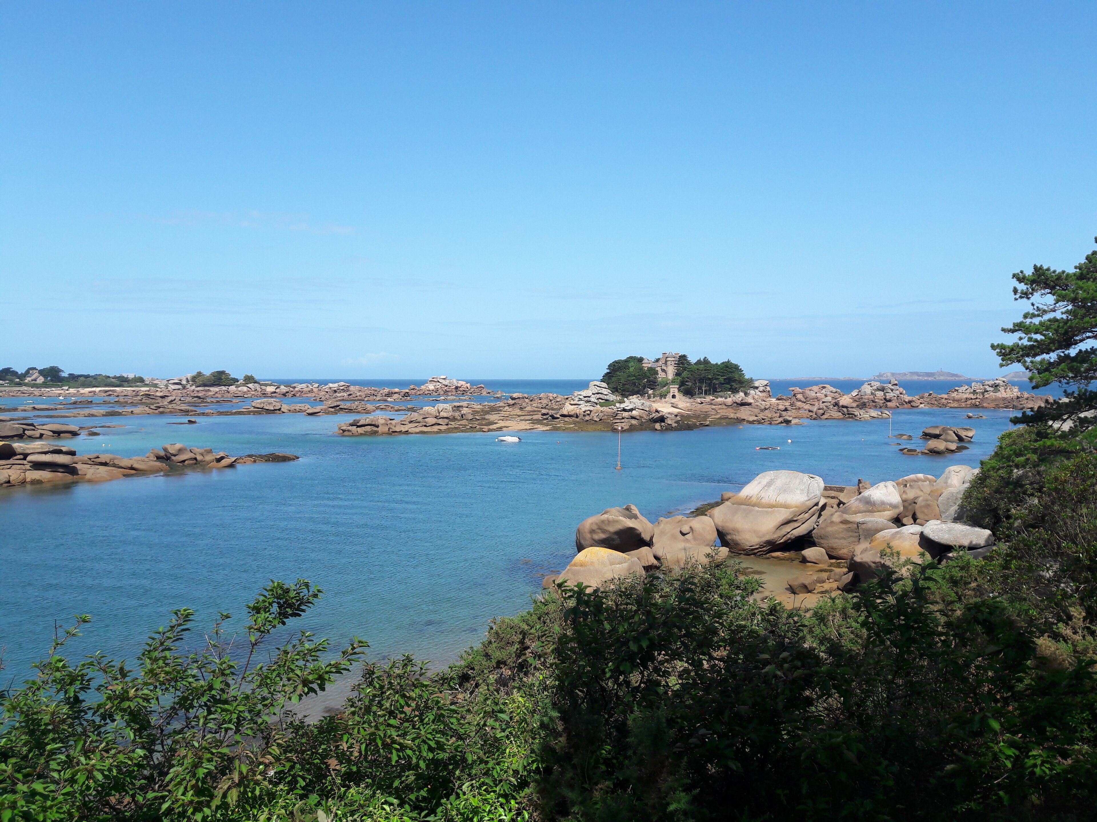 A coastal landscape with rocky outcrops, clear blue water, and a distant building on a hill.