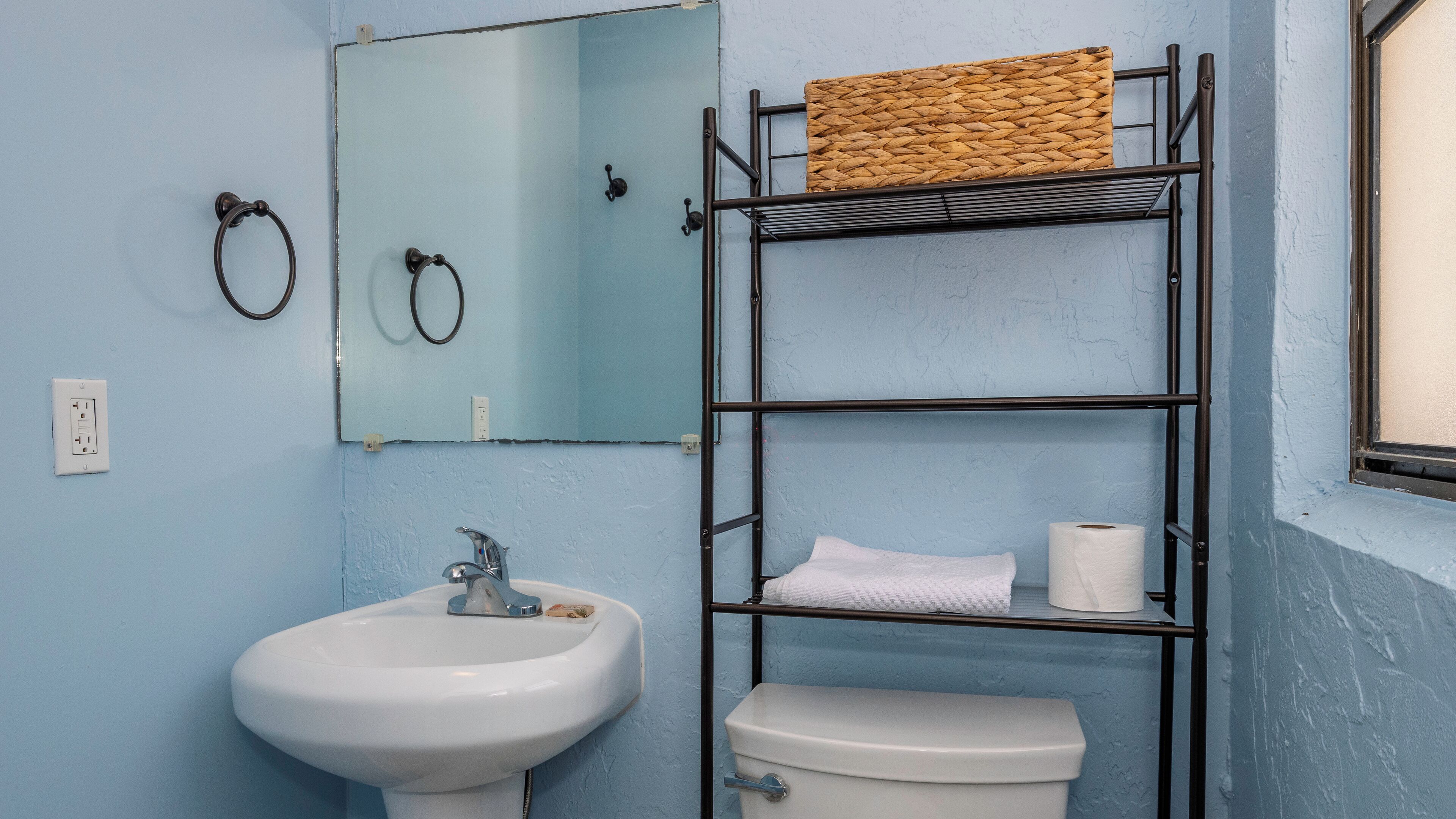 Ein Badezimmer mit einem weißen Waschbecken, einer Toilette und einem Metallregal mit einem gewebten Korb.
