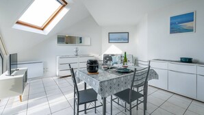 Une cuisine moderne avec un coin repas, des meubles blancs, une verrière et un tableau représentant une scène de plage.