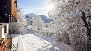 Un sentier enneigé, avec un bâtiment en bois sur la gauche et des arbres chargés de neige sur la droite, qui mène à un paysage montagneux en arrière-plan.