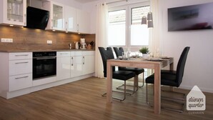 Une cuisine moderne avec des meubles blancs, un four encastré et un coin repas comprenant une table en bois et des chaises noires.
