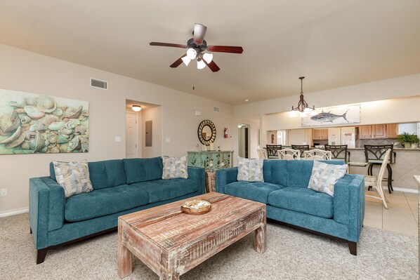 A living room with two teal sofas, a wooden coffee table, a ceiling fan, and a kitchen area in the background. - Waterfront Condo | Pool Access & Stunning Views (Corpus Christi)