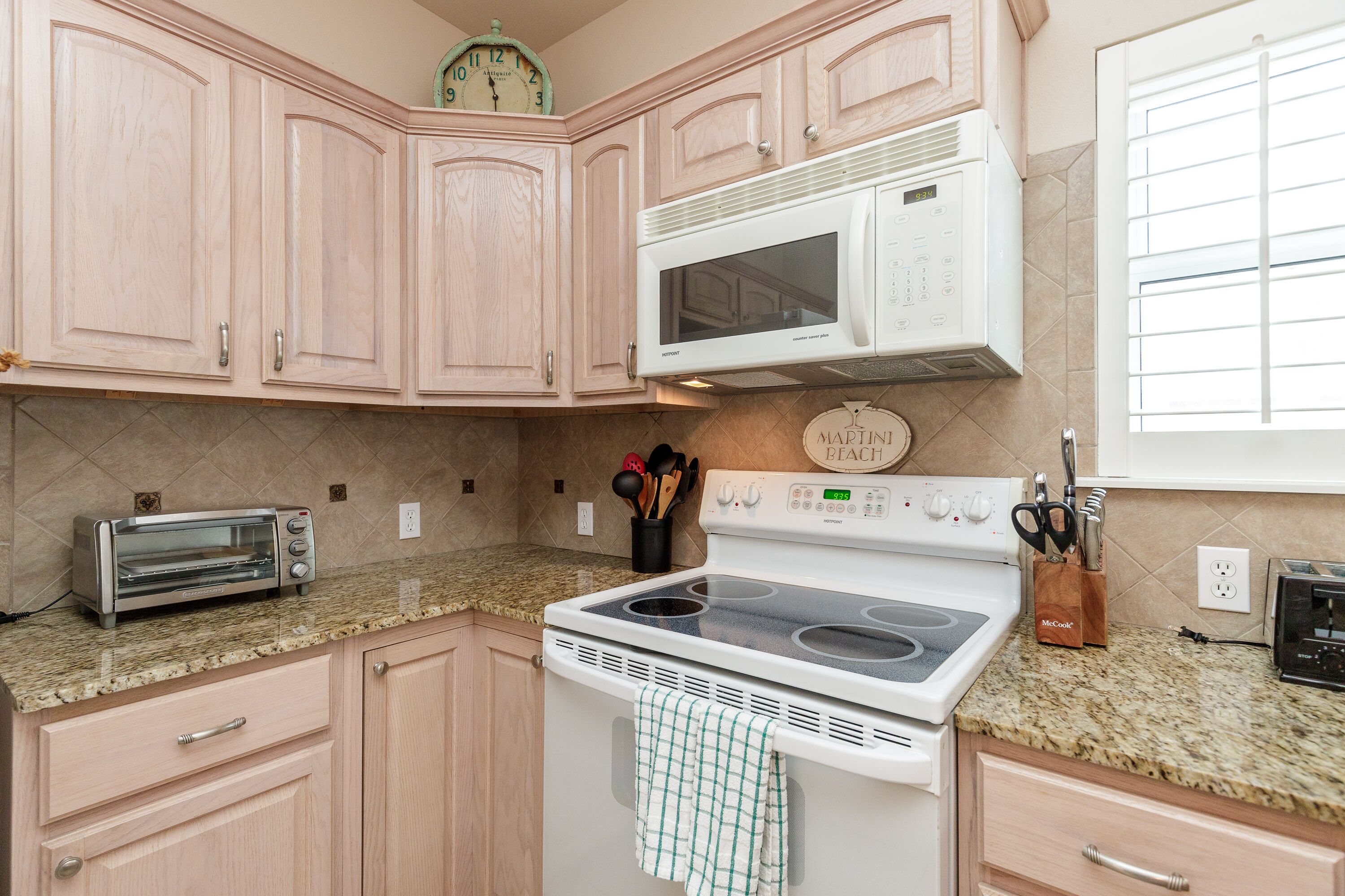 A kitchen with light pink cabinets, a white microwave, a stove, a toaster, and a window with blinds.