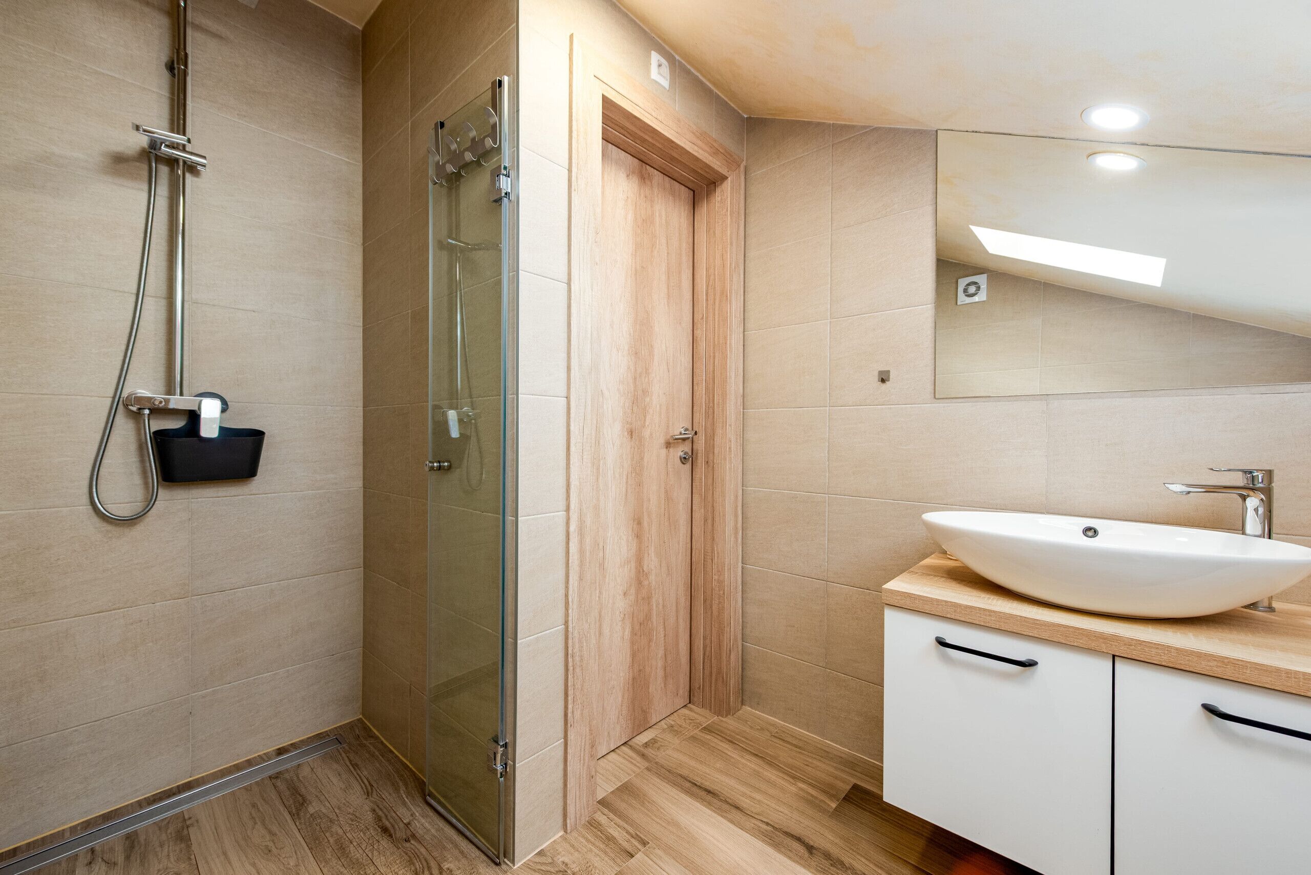 Un baño moderno con cabina de ducha de vidrio, puerta de madera, lavamanos blanco y espejo sobre el lavamanos.