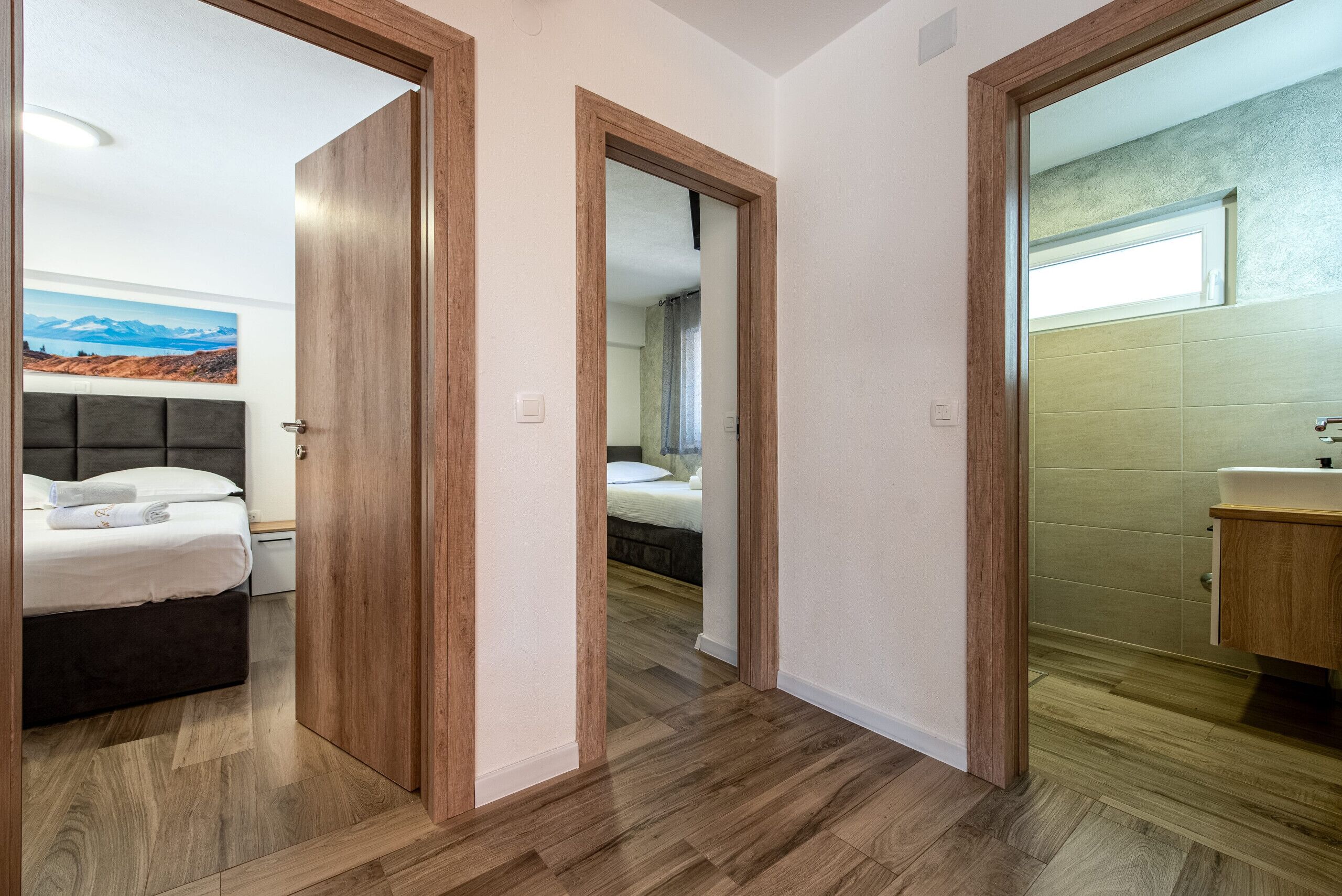 A hotel room with a bed, a bathroom with a sink, and a hallway with wooden flooring and doors.