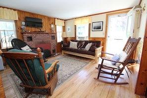 A cozy living room with a fireplace, wooden walls, a sofa, a rocking chair, and a television.