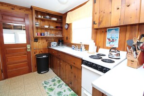 A kitchen with wooden cabinets, a stove, a trash can, and various kitchen items. - Cozy Cottage Near Rock Harbor (2223) (Orleans)