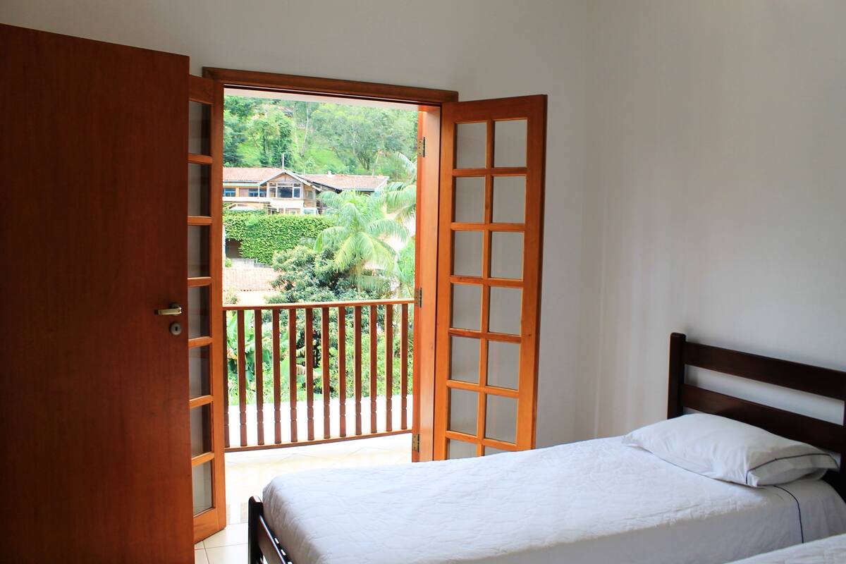 Een slaapkamer met een bed, een houten deur en een balkon met uitzicht op groen en een huis.