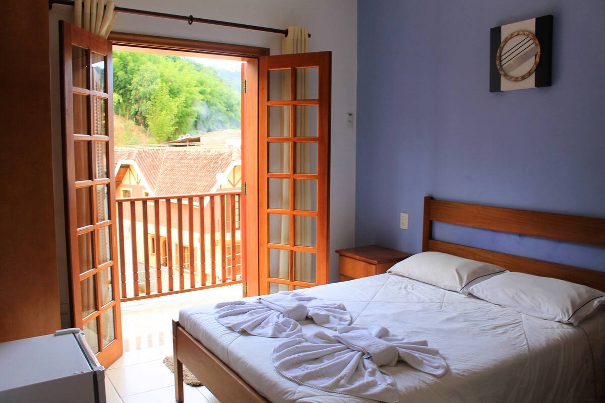 Een slaapkamer met een bed, een houten hoofdbord, een nachtkastje en een raam met uitzicht op een balkon en bomen.