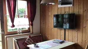 Et hyggeligt værelse med træpaneler, et bord med hvid dug, to stole, en bænk med rød pude, en hængende lampe og et fjernsyn monteret på væggen.
