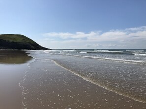 A beach with gentle waves, a sandy shore, and a grassy hill in the background. - Yazelle town house (Llanelli)