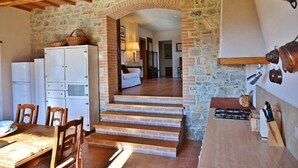 Eine rustikale Küche mit Steinwänden, Holzbalken, einer Essecke mit Stühlen und einer Treppe, die in einen Wohnbereich führt.