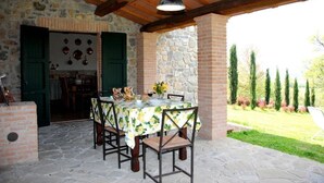Eine Terrasse im Freien mit einem Steintisch und Stühlen, einer hängenden Leuchte und Blick auf einen Garten mit Bäumen und Sträuchern.