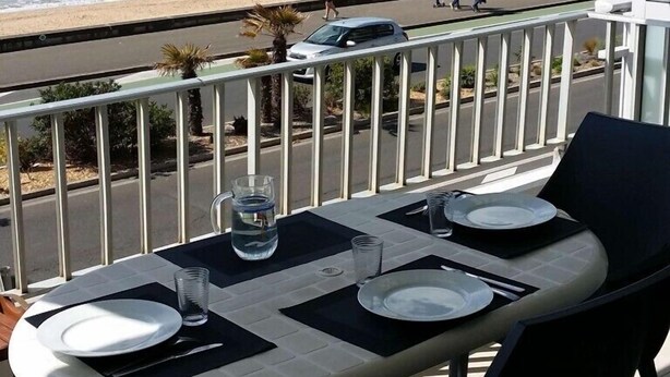 A balcony with a table set for two, overlooking a beach with people walking and a car parked on the road.