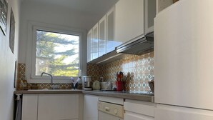 Una cocina compacta con armarios blancos, una ventana con vistas a árboles, una cafetera y un recipiente rojo con lunares.
