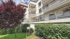 Un immeuble résidentiel à plusieurs étages, doté de balcons, d’un espace jardin agrémenté d’un panneau et d’un arbre aux feuilles rouges.