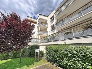 A multi-story residential building with balconies, a garden area with a sign, and a tree with red leaves.