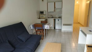 Ein kompakter Wohnbereich mit einem dunkelgrauen Sofa, einem kleinen Holztisch, einer Küchenzeile mit Mikrowelle und Kühlschrank sowie einem Essbereich mit Tisch und Stühlen.