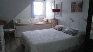 A bedroom with a large bed, a smaller bed, a chair, a nightstand, a lamp, and a window with curtains.