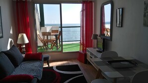 A room with a sofa, a TV, a table with a laptop, a chair, and a view of the sea through large windows.