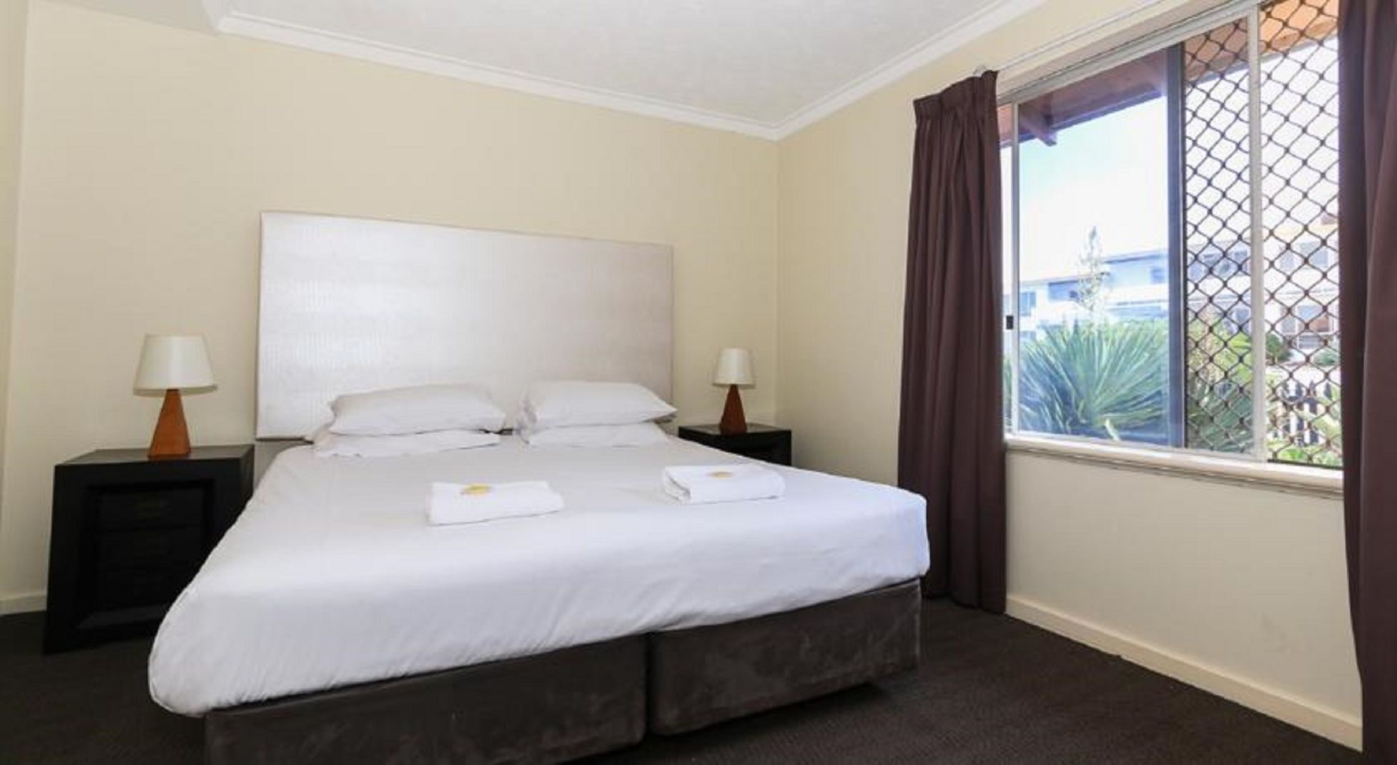 A neatly made bed with white linens and two pillows, a bedside table with a lamp, and a window with curtains.